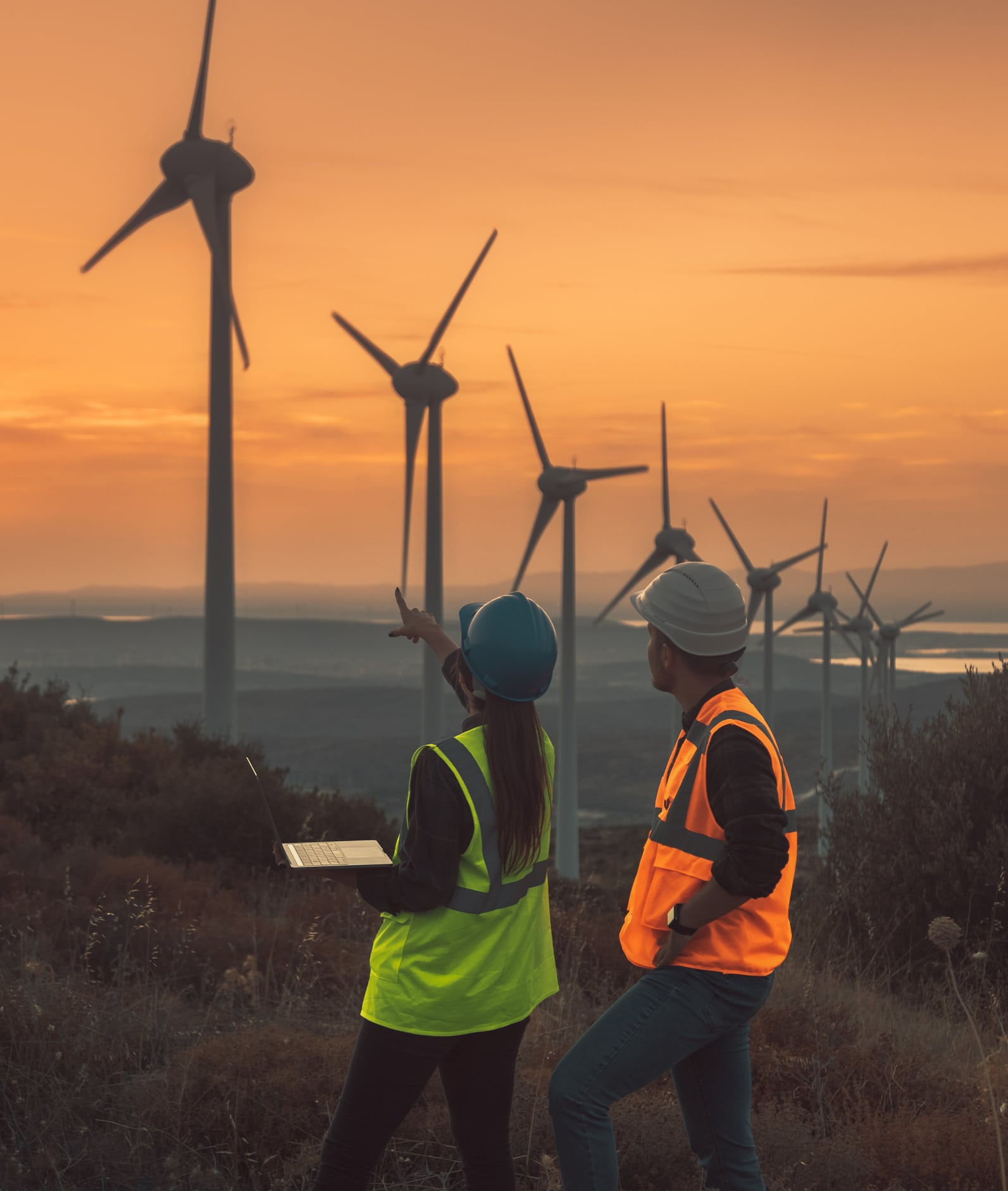 Young electrical engineer woman and business man standing in front of wind turbines checking and working about technical problems and writes the results of measurements with laptop pc in wind power plant electric energy station. xxxl size taken with canon 5d mIV